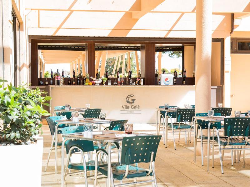 Helle Cafeteria mit grünen Stühlen, weißen Tischen und einer langen Bar im Hintergrund.