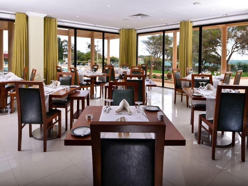 Helles Restaurant mit großen Fenstern, mehreren Holztischen und schwarzen Stühlen mit grünen Vorhängen.