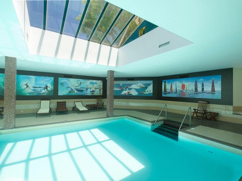 Helles Hallenbad mit Glasdach, breiten Fensterbildern und Sitzgelegenheiten an den Wänden.