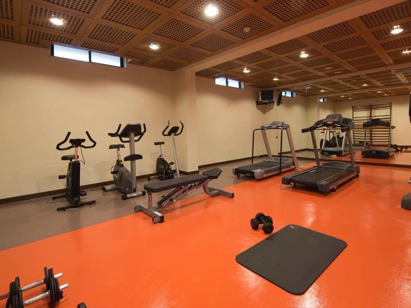 Kleiner Fitnessraum mit oranger Boden, Laufbändern, Fahrrädern, Hanteln und Matte.