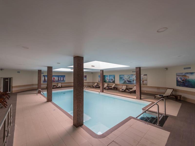 Hallenbad mit quadratischem Pool, hellen Wänden, mehreren Liegestühlen und einer gemalten Unterwasserlandschaft.