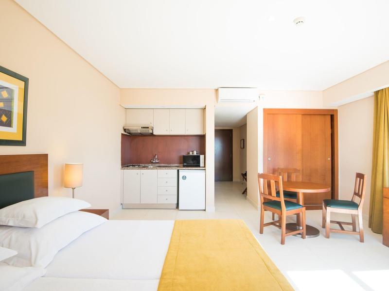 Helles Hotelzimmer mit großem Bett, gelber Decke, Küchenzeile, Tisch, Stühlen und großem Fenster.