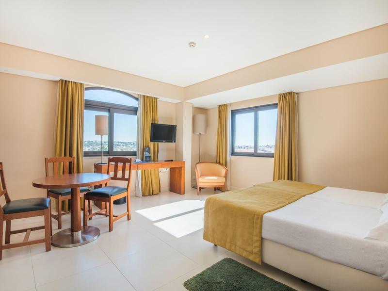 Helles Hotelzimmer mit Doppelbett, Tisch, Stühlen und großen Fenstern mit Ausblick.