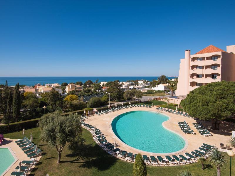 Hotel mit rundem Pool, Sonnenliegen, Bäumen, Häusern und blauem Himmel am Meer im Hintergrund.