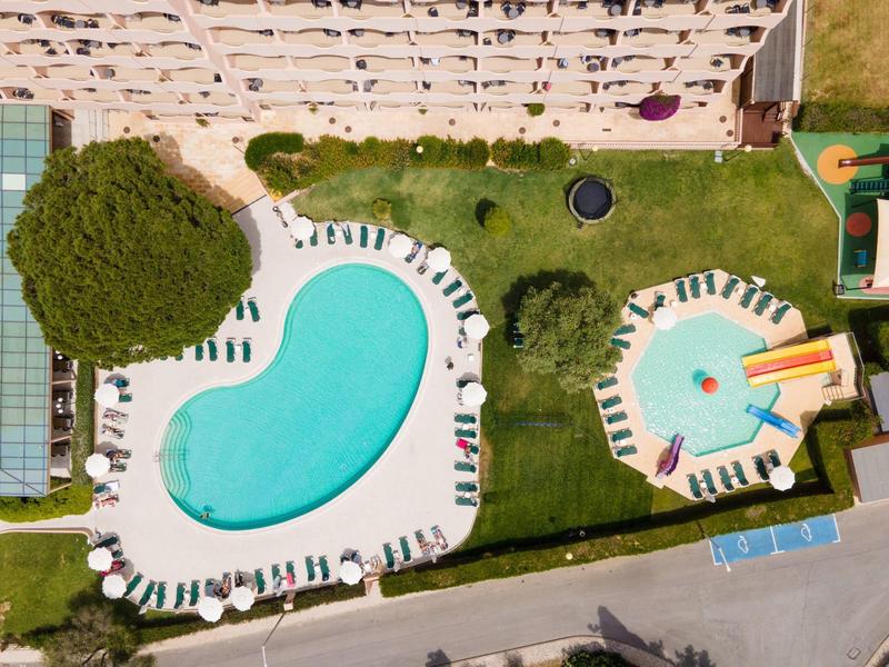 Zwei Hotelpools mit Liegestühlen auf grünem Rasen neben einem Gebäude.