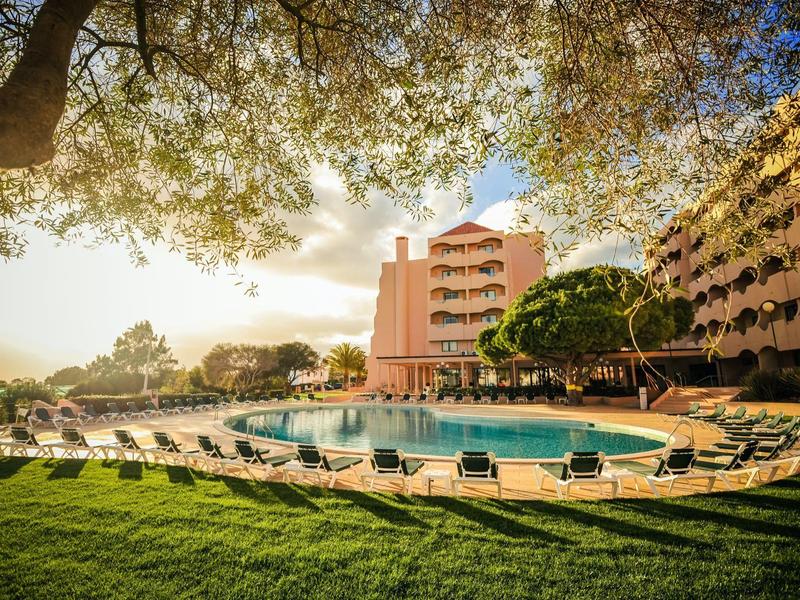 Luxuriöses Hotel mit Außenpool, umgeben von Liegestühlen und grüner Wiese unter Bäumen.