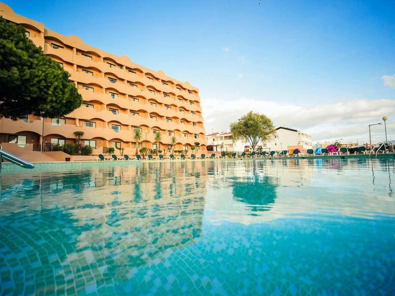 Großes Hotel mit Balkonwohnungen neben einem blauen Pool unter klarem Himmel.