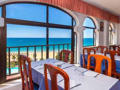 Restaurant mit Holztischen und Stühlen, großen Fenstern und Blick auf einen Strand und das Meer.