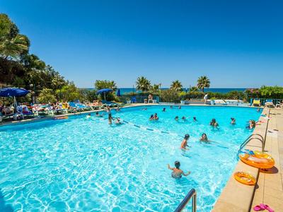 Groot buitenzwembad met mensen en een heldere blauwe lucht, omgeven door palmbomen en ligstoelen.