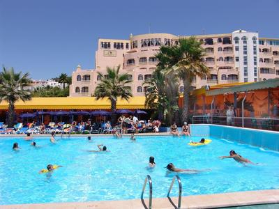 Groot hotel met een zwembad waar gasten zwemmen en ontspannen onder een heldere hemel.