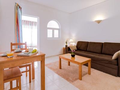 Lichte woonkamer met bruine bank, houten salontafel en eettafel naast een raam met kleurrijke gordijnen.