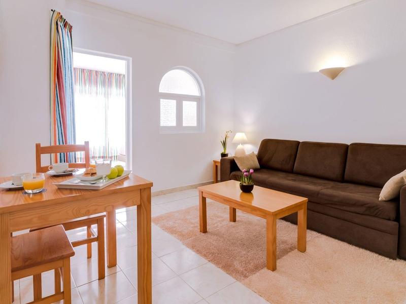 Lichte woonkamer met bruine bank, houten salontafel en eettafel naast een raam met kleurrijke gordijnen.