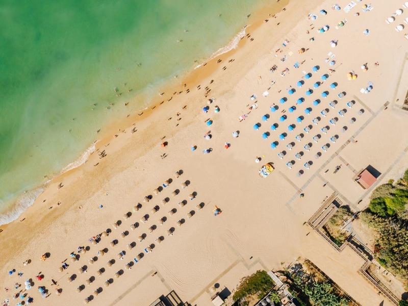Luchtfoto van een strand met parasols, groen water en aangrenzende gebouwen.