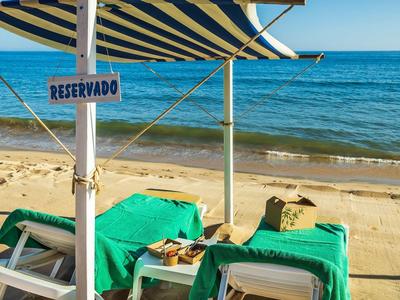 Dva lehátka se zelenými ručníky pod modro-bílým pruhovaným slunečníkem na pláži s cedulkou rezervováno.