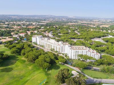 Velká budova hotelu obklopená zelenými stromy a golfovým hřištěm pod jasnou oblohou.