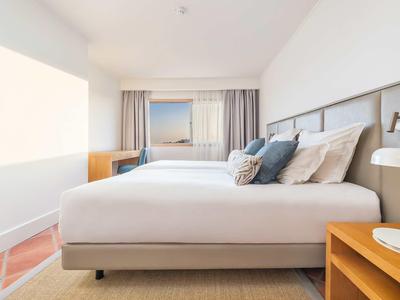 Modernes Hotelzimmer mit großem Bett, Kissen und Blick auf ein Meer durch das Fenster.