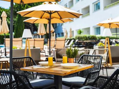 Outdoor-Restaurantbereich mit Sonnenschirmen, modernen Stühlen und Holztischen vor einem Hotelgebäude.