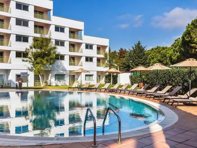 Kruhový venkovní bazén s lehátky a slunečníky před moderní hotelovou budovou.