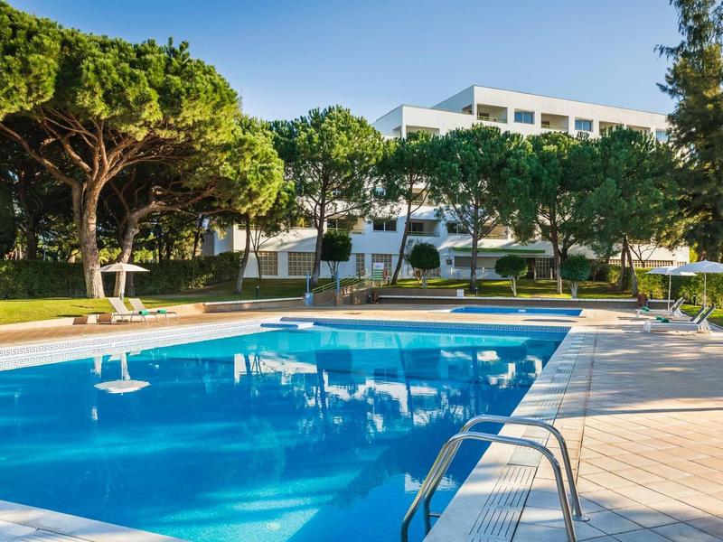 Moderní bazén s čistou modrou vodou vedle bílého hotelového domu a stromů.