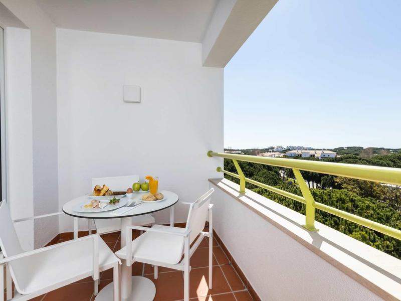 Balkon s bílým stolem a židlemi, výhled na zelenou krajinu pod jasnou oblohou.