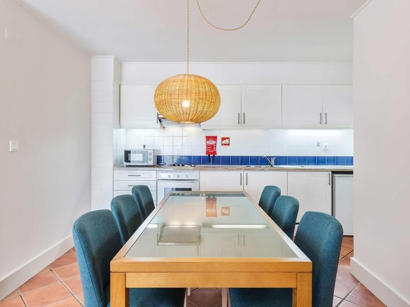 Moderní jídelní prostor s dřevěným stolem, modrými židlemi a otevřenou kuchyní v pozadí.