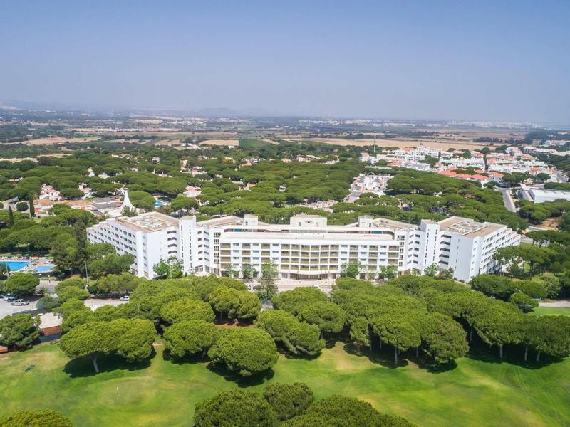 Velká bílá hotelová budova obklopená zelenými stromy a golfovým hřištěm pod jasnou oblohou.