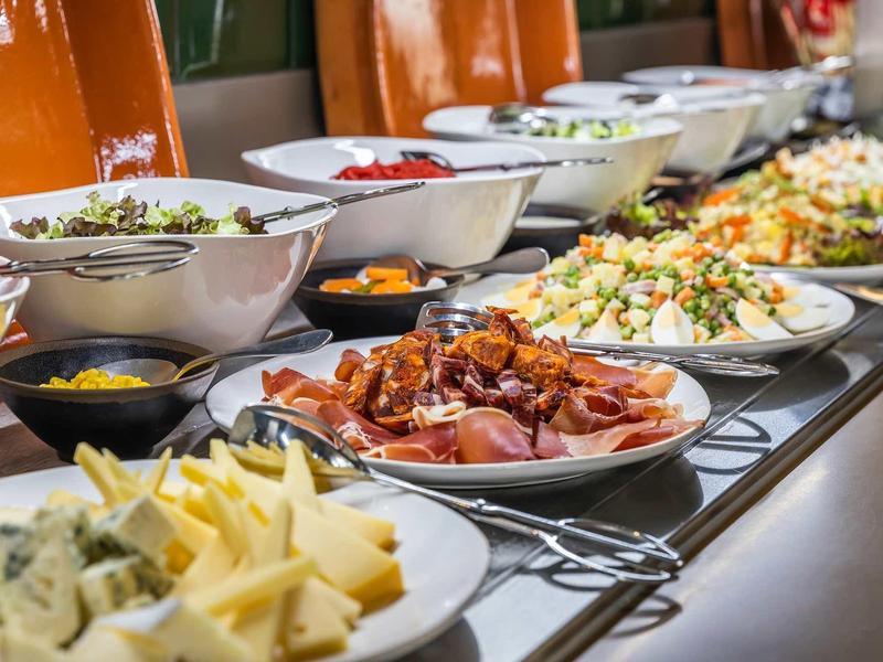Bufet s různými jídly, včetně sýrů, šunky a salátů v miskách a na talířích.