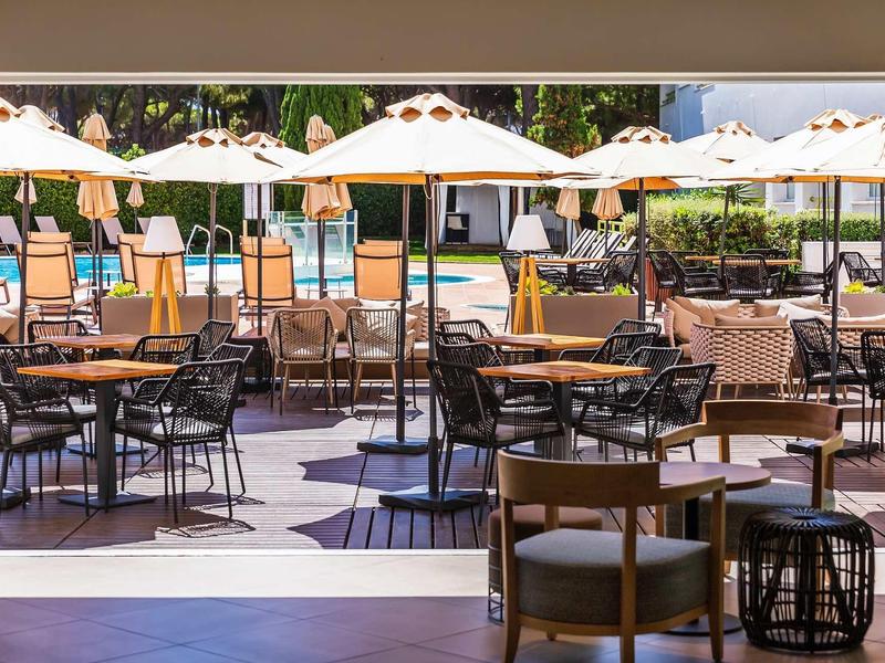 Venkovní část kavárny s stoly, židlemi a bílými slunečníky během dne.