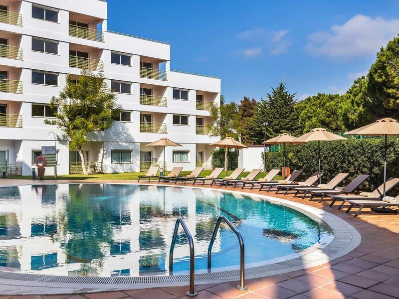 Kruhový venkovní bazén s lehátky a slunečníky před moderní hotelovou budovou.