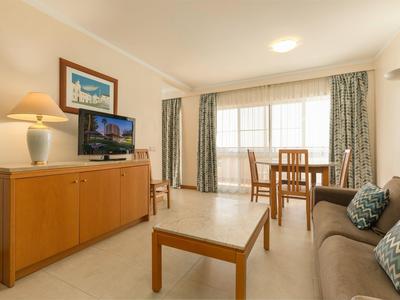 Sala de estar acogedora con sofá, mesa, televisor y comedor frente a grandes ventanas con cortinas.