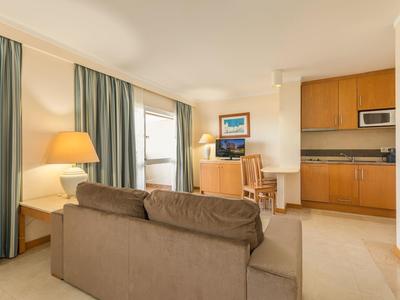 Sala luminosa con sofá marrón, escritorio, cocina pequeña y ventana con cortinas verdes.