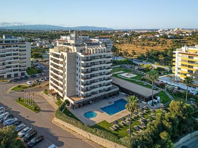 Edificio de hotel moderno con piscina, rodeado de áreas verdes y estacionamiento en zona urbana.