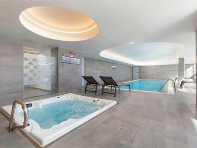 Área interior moderna con jacuzzi, piscina y tumbonas bajo techo blanco.