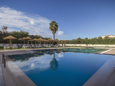 Großer Außenpool mit blauer Wasserfläche, umgeben von Liegestühlen und Sonnenschirmen unter blauem Himmel.