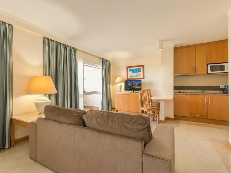 Sala luminosa con sofá marrón, escritorio, cocina pequeña y ventana con cortinas verdes.