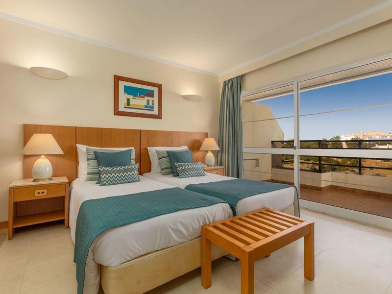 Habitación de hotel luminosa con dos camas individuales, detalles azules y balcón con vista.