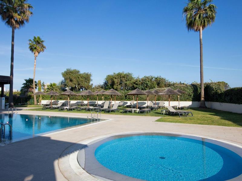 Área de piscina con tumbonas, dos piscinas circulares y palmeras bajo cielo despejado.