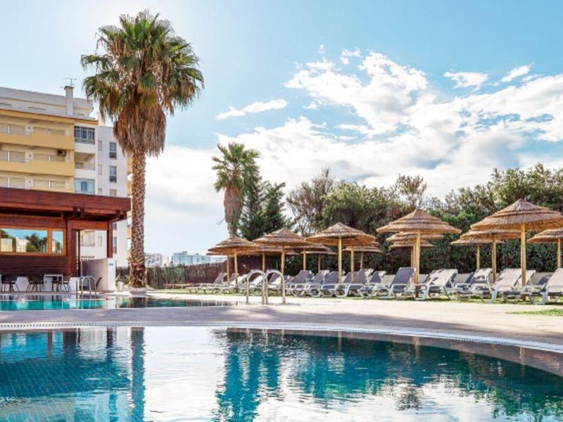 Gran piscina al aire libre con tumbonas y sombrillas frente a edificio del hotel en día soleado.