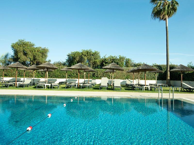 Piscina clara con tumbonas y sombrillas en un jardín soleado de hotel con palmeras.
