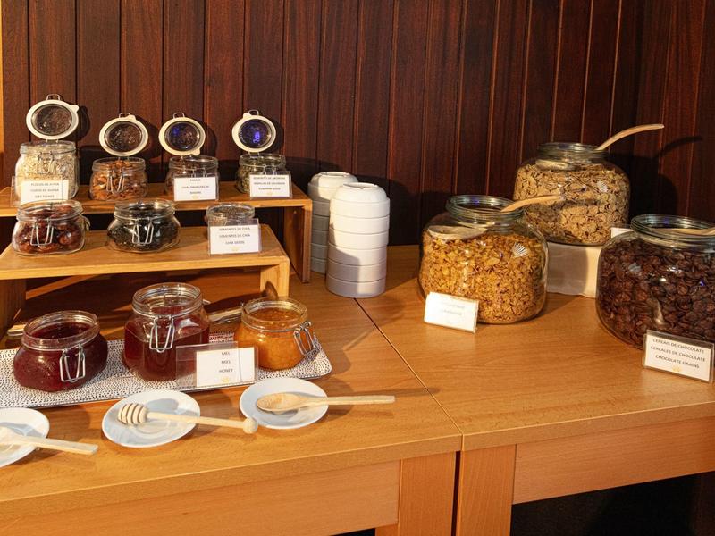Mesa con frascos de mermeladas y cereales en un buffet de desayuno de hotel.