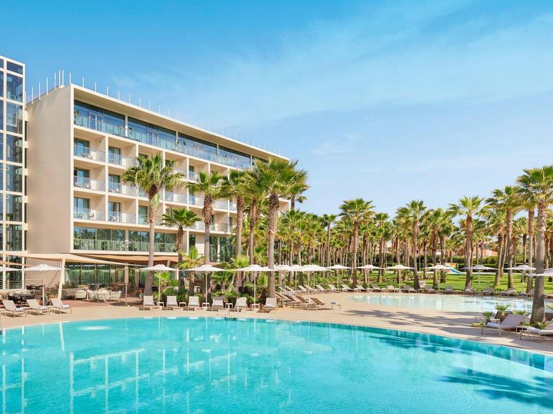 Hôtel moderne avec grande piscine et palmiers sous un ciel bleu.