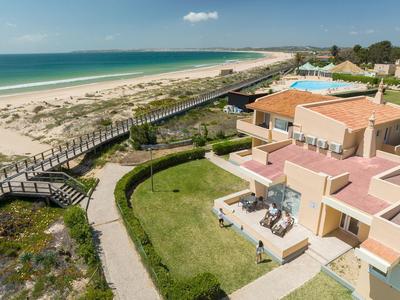 Hotel mit Terrasse, Pool und Sandstrand an der türkisfarbenen Küste unter blauem Himmel.