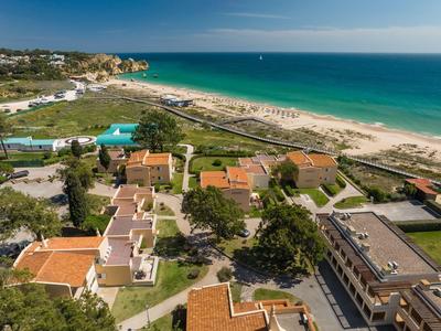 Luftaufnahme eines Strandes mit angrenzenden Gebäuden und grünen Flächen unter blauem Himmel.