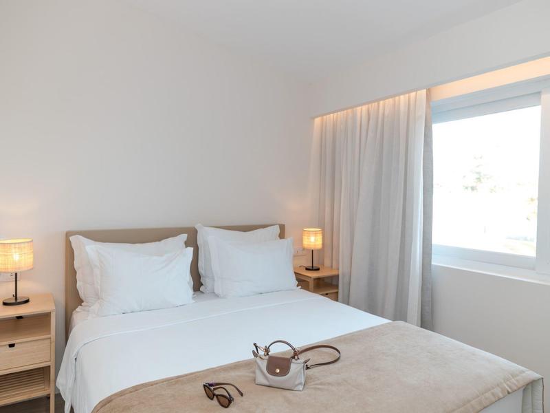 Hell eingerichtetes Schlafzimmer mit Doppelbett, Nachtlampen und Fenster mit Vorhängen.