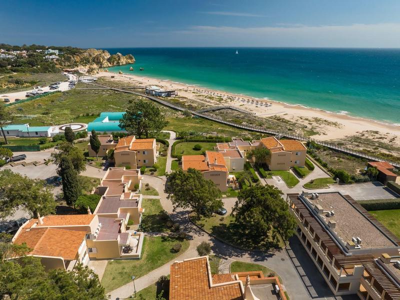Luftaufnahme eines Strandes mit angrenzenden Gebäuden und grünen Flächen unter blauem Himmel.