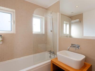 Helles Badezimmer mit Badewanne, Glasdusche, Waschbecken auf Holzunterschrank und zwei Fenstern.