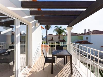 Balkon mit Tisch und zwei Stühlen, überdacht mit dunklen Holzbalken, in sonniger Umgebung.