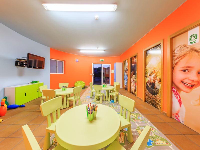 Bunter Kinderspielraum mit kleinen Tischen, Stühlen, Spielzeug und großen Bilderpostern an der Wand.