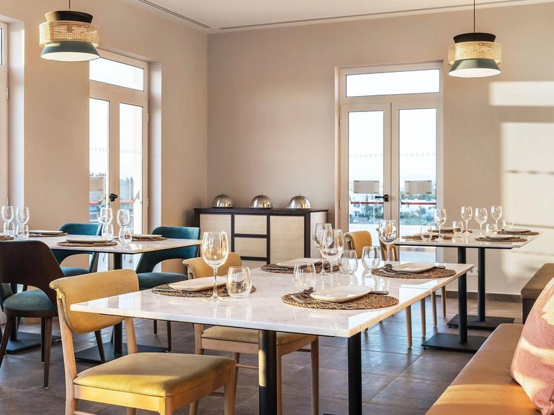 Moderner, heller Speiseraum mit weißen Tischen, bequemen Stühlen und großen Fenstern für Tageslicht.