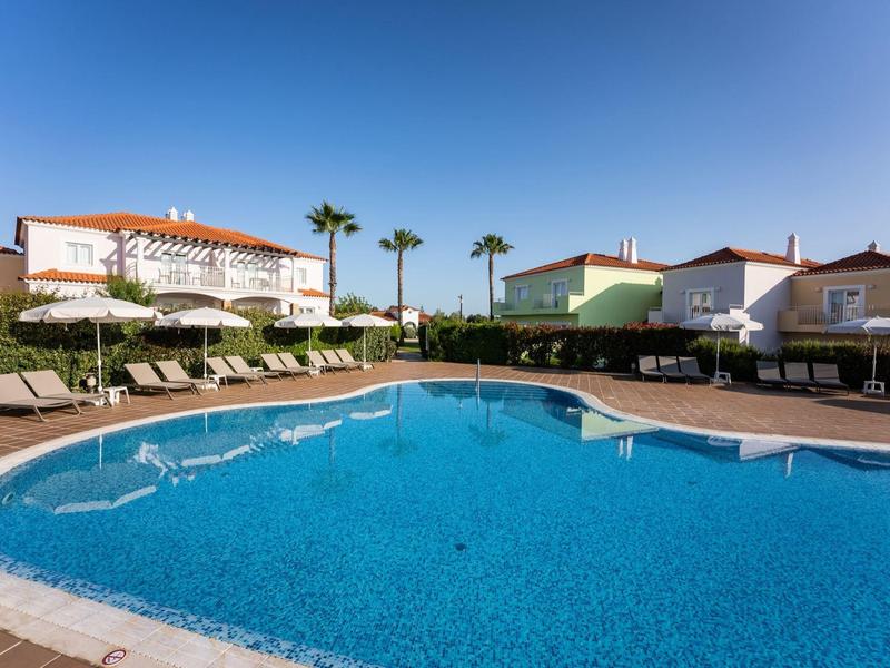 Großer blauer Pool mit Sonnenliegen, Palmen und weißen Häusern unter klarem Himmel.